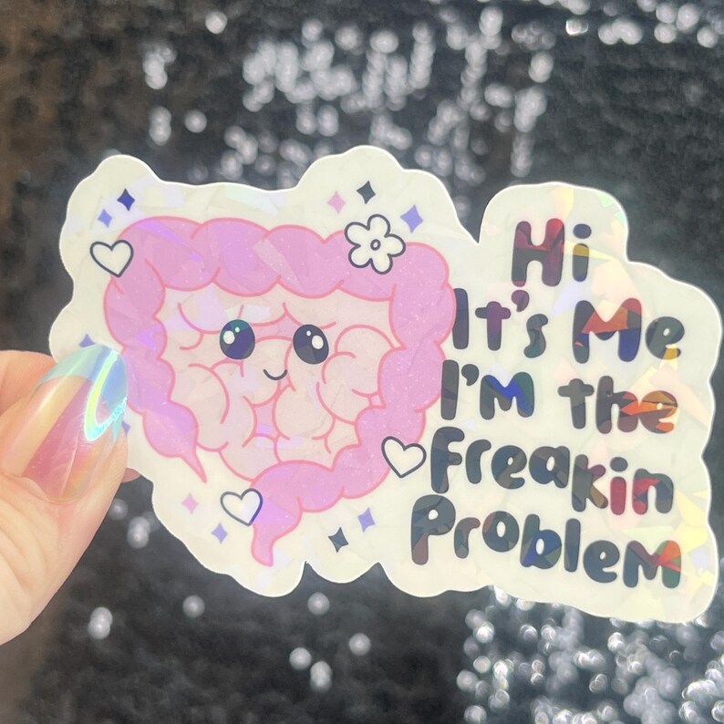 May include: A holographic sticker featuring a cartoon intestine with a smiling face, a flower, and hearts. The sticker has the text "Hi It's Me I'm the Freakin Problem" in colorful letters. The sticker is held by a hand.