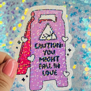 May include: A holographic sticker in the shape of a caution sign, with the text "CAUTION: YOU MIGHT FALL IN LOVE." The sign has a pink and purple glittery background, with a graphic of a person falling in love.