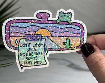 May include: A holographic sticker with a rearview mirror design. The mirror shows a sunset over a desert landscape with a cactus. A note attached to the mirror says "Don't look back, you're not going that way". A frog sits on top of the mirror.