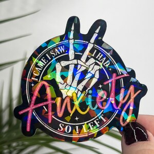 K&ouml;nnte beinhalten: Ein holografischer Aufkleber mit einem Peace-Zeichen, das aus einer Skeletthand besteht. Der Aufkleber zeigt den Text "I came I saw I had anxiety so I left" um das Peace-Zeichen herum.