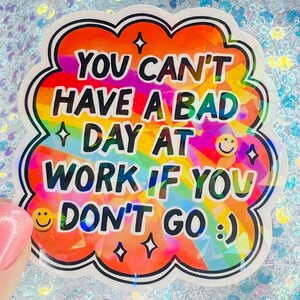 Puede incluir: Una pegatina colorida con el texto "YOU CAN'T HAVE A BAD DAY AT WORK IF YOU DON'T GO :)". La pegatina tiene un fondo de arco&iacute;ris, un contorno blanco y un borde negro. Caritas sonrientes y estrellas tambi&eacute;n est&aacute;n incluidas en el dise&ntilde;o.