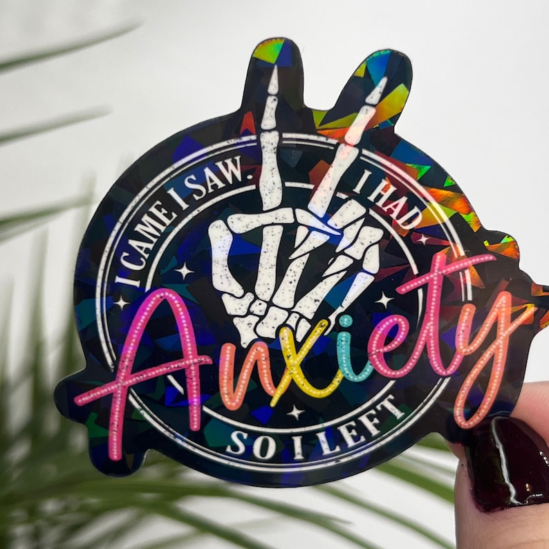 K&ouml;nnte beinhalten: Ein schwarz-wei&szlig;er Aufkleber mit einer Skeletthand, die ein Peace-Zeichen macht. Der Aufkleber hat einen regenbogenfarbenen holografischen Hintergrund und den Text "I came I saw I had anxiety so I left".
