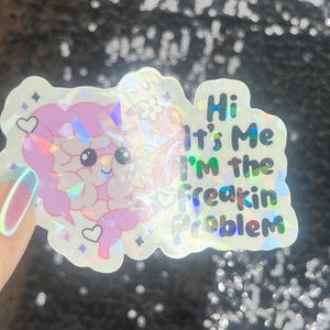 May include: A holographic sticker with a cartoon brain design in pink and purple. The sticker has the text "Hi It's Me I'm the Freakin Problem" in green and blue. The sticker is decorated with hearts and stars.