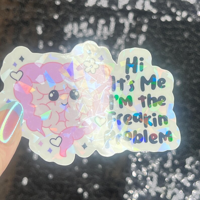 May include: A holographic sticker with a cartoon brain design in pink and purple. The sticker has the text "Hi It's Me I'm the Freakin Problem" in green and blue. The sticker is decorated with hearts and stars.