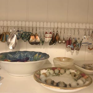 Könnte beinhalten: Ein weißer Kunststoff-Ohrringhalter mit einer Reihe von Haken. An den Haken hängen verschiedene Ohrringe, darunter Schmetterlings-, Blumen- und geometrische Designs. Die Ohrringe sind aus Metall, Glas und Kunststoff.