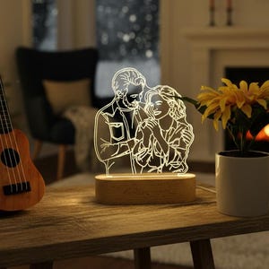 Könnte beinhalten: Eine leuchtende LED-Lampe mit einem sich umarmenden Paar, auf einem Holzsockel. Die Lampe steht auf einem kleinen Holztisch, zusammen mit einer Ukulele, einer Topfpflanze mit gelben Blumen und einem Weihnachtsbaum im Hintergrund. Gemütliches Wohnzimmer.