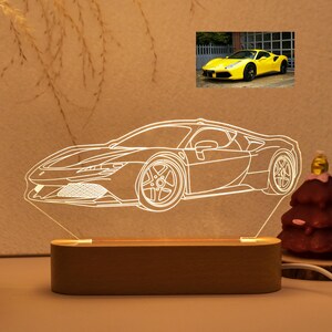 Könnte beinhalten: Eine LED-Lampe in Form eines Autos mit Holzsockel. Die Lampe strahlt ein helles, weißes Licht aus und zeigt die Konturen eines Sportwagens. Ein kleines Bild eines gelben Sportwagens befindet sich im Hintergrund. Rechts eine Weihnachtsbaumdekoration.