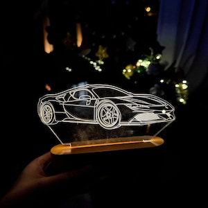 May include: A clear acrylic car-shaped night light with a wooden base. The car is outlined in white, showcasing the vehicle's design. The light is held in a hand against a dark background.