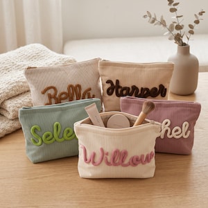 Könnte beinhalten: Fünf Reißverschlusstaschen aus Kord in verschiedenen Farben wie Beige, Salbeigrün und Mauve. Jede Tasche ist auf der Vorderseite mit einem Namen in erhabener, strukturierter Schrift bestickt. Eine offene Tasche enthält Lippenbalsam und einen Pinsel.