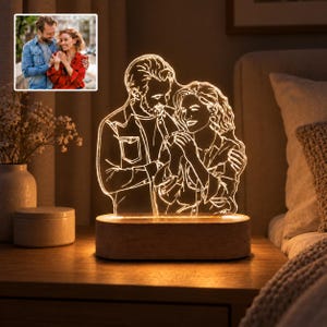 Może przedstawiać: Personalizowana lampka nocna LED z podświetlanym, grawerowanym rysunkiem liniowym przedstawiającym przytulającą się parę, na podstawie załączonego zdjęcia. Panel akrylowy jest osadzony na prostokątnej drewnianej podstawie emitującej bursztynowe światło.