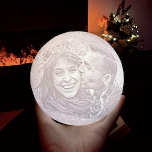 May include: A spherical moon lamp featuring a 3D-printed relief portrait of two people, one kissing the other on the cheek. The surface of the lamp has a textured, cratered appearance resembling the moon, and it emits a soft white glow.