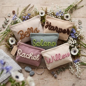 Könnte beinhalten: Fünf rechteckige Reißverschlusstaschen aus geripptem Stoff in Beige, Salbeigrün und Altrosa. Jede Tasche ist mit einem personalisierten Namen in erhabener Schreibschrift versehen: Bella, Harper, Selena, Rachel und Willow, inklusive kleinem Anhänger.