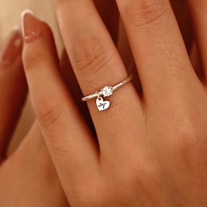 Könnte beinhalten: Ein silberner Ring mit einem kleinen, klaren Edelstein und einem herzförmigen Anhänger. Der Anhänger trägt den Buchstaben "A". Der Ring befindet sich an einem Finger.