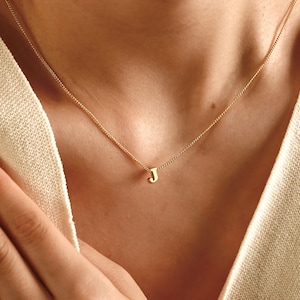 Könnte beinhalten: Eine zarte goldene Halskette mit einem kleinen Anhänger in Form des Buchstabens "J". Der Anhänger hängt an einer dünnen Goldkette. Die Halskette wird um den Hals getragen und liegt auf der Brust.