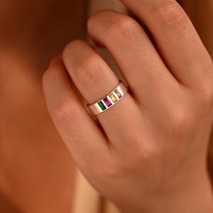 Könnte beinhalten: Ein silberner Ring mit vier rechteckigen Edelsteinen in Grün, Rosa, Gelb und Orange. Der Ring befindet sich an einem Finger, die Edelsteine sind in einer Reihe angeordnet.