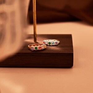 Könnte beinhalten: Zwei Ringe mit farbigen Edelsteinen, die auf einem dunklen Holzblock ausgestellt sind. Ein Ring ist goldfarben mit einer Reihe von rosa, grünen, orangefarbenen und lila Steinen. Der andere Ring ist silberfarben mit grünen, blauen und lila Steinen.