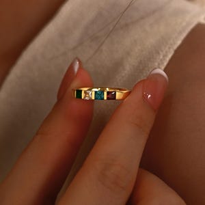 Könnte beinhalten: Ein goldener Ring mit fünf quadratischen Edelsteinen in Grün, Weiß, Türkis und Rot. Der Ring wird zwischen Fingern gehalten, wodurch die farbigen Steine und das Goldband hervorgehoben werden.