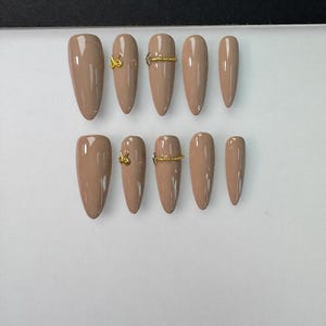 May include: A set of ten almond-shaped press-on nails in a glossy nude beige finish. Some nails feature small gold-toned metal embellishments, including tiny bow-shaped charms and thin horizontal bands that resemble rings across the nail surface.