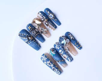 May include: A set of long, coffin-shaped artificial nails. The nails feature a denim-like blue color, adorned with intricate designs, rhinestones, and gold accents. Some nails have a nude base with embellishments.