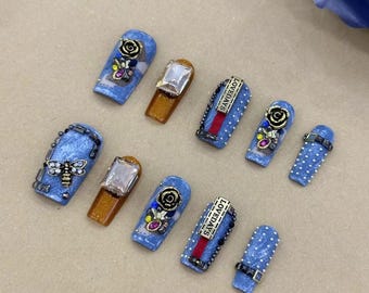 Puede incluir: Un conjunto de diez uñas postizas azules y blancas con varios adornos, incluyendo piedras de imitación, un amuleto de abeja y una barra dorada con el texto "Lovedays".
