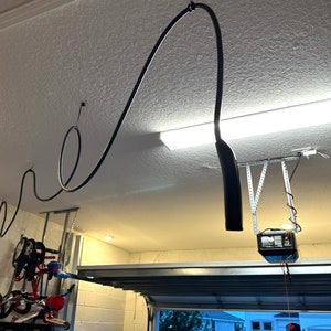 Puede incluir: Un cable eléctrico negro cuelga del techo de un garaje. El cable está enrollado y corre a lo largo del techo. Se puede ver un abrepuertas de garaje en el fondo.