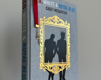 May include: A hardcover book titled "RED, WHITE & ROYAL BLUE" by Casey McQuiston. The book has a gray cover with gold accents and a gold-framed silhouette of two figures. A red ribbon bookmark with a crown charm is attached.