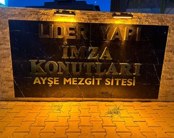 May include: A dark marble sign with gold lettering reads "LIDER YAPI IMZA KONUTLARI AYSE MEZGIT SITESI". The sign is illuminated by overhead lights and set against a brick wall. The scene is lit at night.