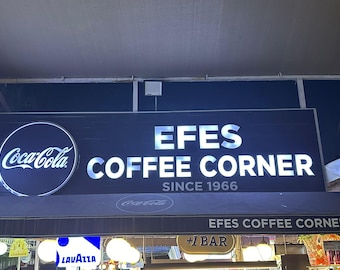 May include: A black and white sign with white lettering that reads "EFES COFFEE CORNER SINCE 1966". A Coca-Cola logo is on the left side of the sign.