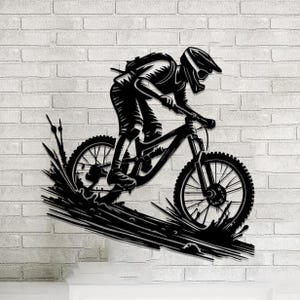 May include: Black metal wall art depicting a mountain biker in action. The cyclist is wearing a helmet and riding a mountain bike, with water splashing up from the tires. The artwork is mounted on a white brick wall.