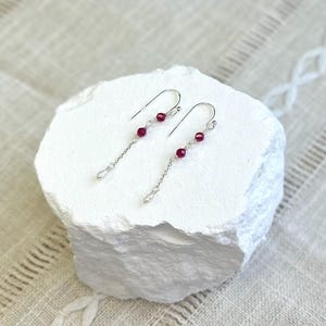 Könnte beinhalten: Ein Paar silberne Ohrhänger mit jeweils zwei kleinen, facettierten, dunkelroten Perlen an einer dünnen Kette. Die Ohrringe haben geschwungene Ohrhaken und enden unten mit einer winzigen, glatten Silberperle für ein zartes Erscheinungsbild.