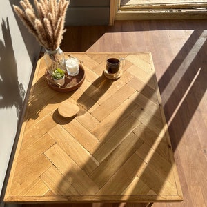 Puede incluir: Una mesa de café de madera con una parte superior de patrón de espiga. La mesa está hecha de madera marrón claro y tiene un acabado natural. La mesa está sentada en un piso de madera de color marrón claro. Hay una taza de café y una pequeña planta en la mesa.