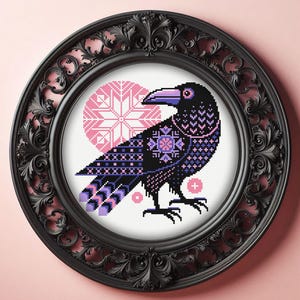 May include: A cross-stitch embroidery of a black raven with purple and pink geometric patterns, set against a pink circular snowflake motif. The artwork is encased in a round, ornate black frame with intricate scrollwork and floral carvings.