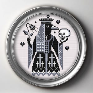 May include: Cross-stitch artwork in a silver frame. The design depicts a crow-like figure wearing a crown and holding a flower and a skull. The colour scheme is black, white, and grey, with cross patterns and gothic elements.