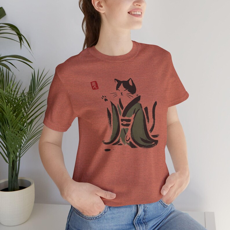 May include: A rust-colored short-sleeve t-shirt featuring a black and olive green ink-wash style illustration of a cat wearing a kimono. The cat is depicted with one paw raised, and a small red square stamp graphic is visible near the cat's head.