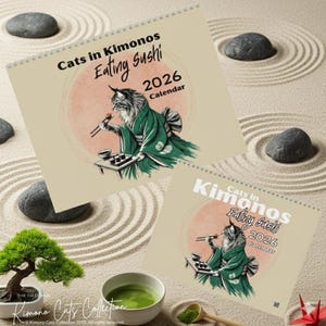 Two spiral-bound wall calendars are displayed on a sand-raked surface, both titled "Cats in Kimonos Eating Sushi 2026 Calendar" with a cat illustration.