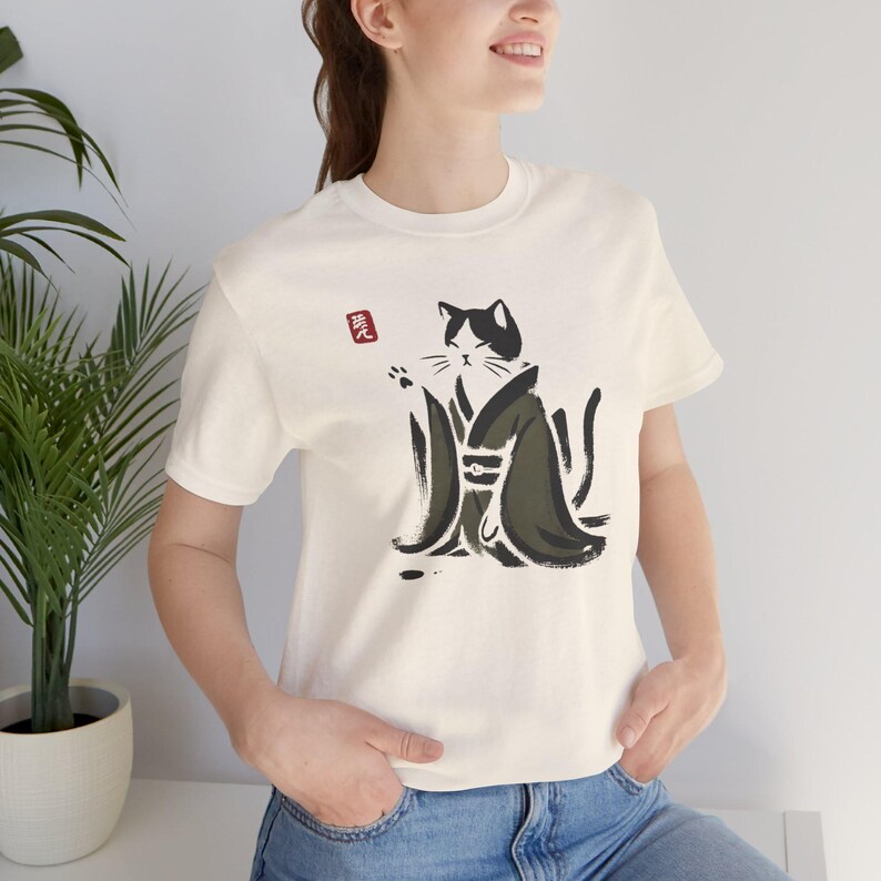 May include: A cream-colored short-sleeve t-shirt featuring a black and white cat illustration wearing a dark green robe. The cat is depicted in a brush-stroke style with one paw raised. A small red square with white Japanese characters is visible above the cat.