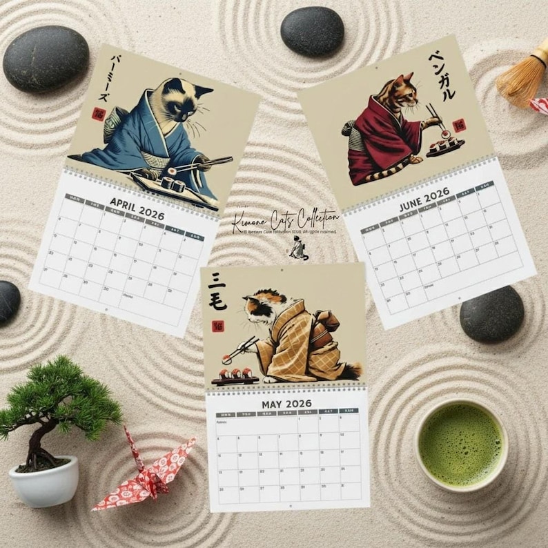 Three calendar pages for April, May, and June 2026 are arranged on a sand-raked surface, each featuring a cat in a kimono eating sushi.