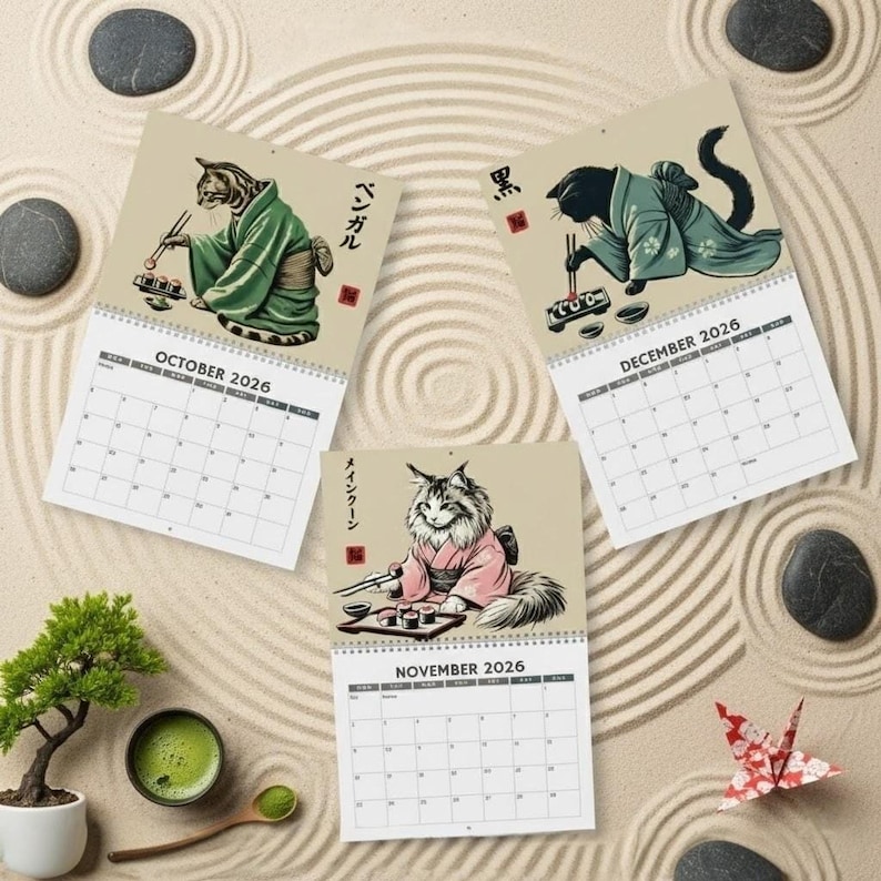 Three calendar pages for October, November, and December 2026 are arranged on a sand-raked surface, each featuring a cat in a kimono eating sushi.