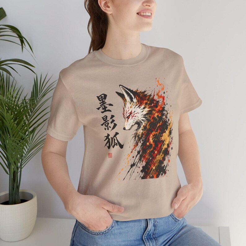 A woman wears a tan t-shirt with a brush stroke fox design in black, orange, and gold, and Japanese characters on the left.