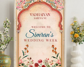 May include: A wedding week announcement sign with a floral design. The sign is framed in a light wood frame and has a peach background. The text on the sign reads "Vadhaiyan Sareya Nu Welcome to Simran's Wedding Week".
