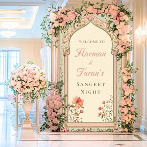 May include: A wedding welcome sign with pink flowers and greenery. The sign says "Welcome to Harman & Taran's Sangeet Night".