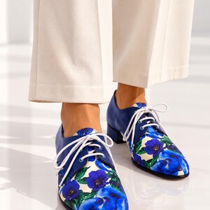 Front view of blue floral leather Oxford shoes worn with ivory trousers.