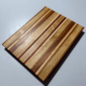 May include: A rectangular wooden chopping board with a striped design. It has alternating light and dark wood strips, with a thin red strip. The board is approximately 30 cm long and 23 cm wide.