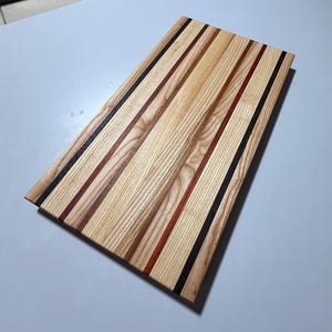 May include: A rectangular wooden chopping board with a light wood grain. The board features vertical stripes of varying widths in light wood, dark brown, red, and black. The chopping board is approximately 46 cm long and 30 cm wide.