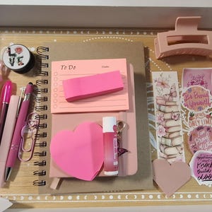 以下が含まれることがあります： ピンクと薄ピンクの文房具のコレクション。ペン、メモ帳、付箋、ヘアクリップ、ステッカーなど。アイテムは装飾的なボーダーが付いた木製のトレイに配置されています。リップグロスとポップソケットも表示されています。