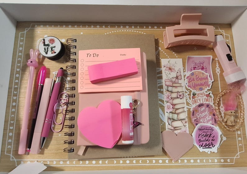 以下が含まれることがあります： ピンクと薄ピンクの文房具のコレクション。ペン、メモ帳、付箋、ヘアクリップ、ステッカーなど。アイテムは装飾的なボーダーが付いた木製のトレイに配置されています。リップグロスとポップソケットも表示されています。