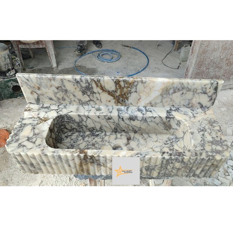 May include: A rectangular marble sink featuring a cream and dark grey veined pattern with a high backsplash and a fluted front design. The basin includes three pre-drilled faucet holes and a central drain opening. A small square logo is visible.