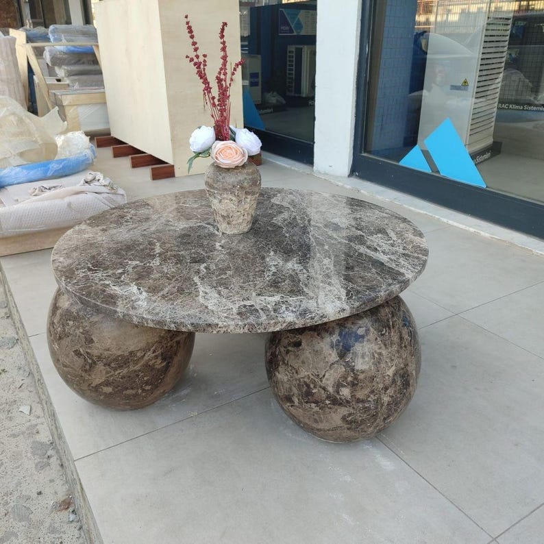 Op de afbeelding: Een ronde marmeren salontafel met een bruin en grijs patroon. De tafel heeft twee grote, bolvormige poten. Op de tafel staat een vaas met bloemen. De vaas heeft een textuur en bevat rode, witte en roze bloemen.