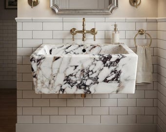 May include: A rectangular wall-mounted bathroom sink crafted from white marble with prominent dark purple and grey veining. The basin features a central drain hole and is paired with a wall-mounted brass faucet fixture and a matching towel ring.