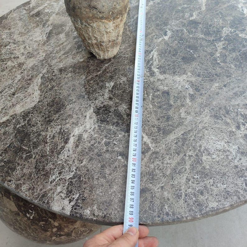 Op de afbeelding: Een ronde, grijze marmeren tafel met een textuur. Een ruw, kegelvormig object staat op de tafel. Een meetlint toont een diameter van ongeveer 90 cm.
